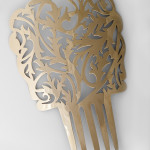 Brass peineta with an elaborate leaf or flame like pattern pierced is photographed on a white background.