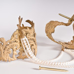 A pair of cast bronze lace handcuffs hinged with fabricated nail closures attached together by a strand of pearls on a white background.