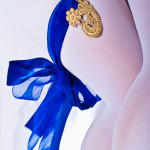 A cast lace piece attached to a garter of royal blue ribbon on female legs wearing white tights on a white background.
